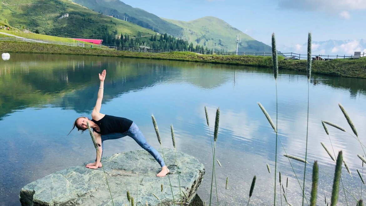 © Rauriser Hochalmbahnen Eine Frau macht Yoga auf einem Felsen am See, im Hintergrund grüne Hügel und blauer Himmel. (vergrößerte Ansicht)