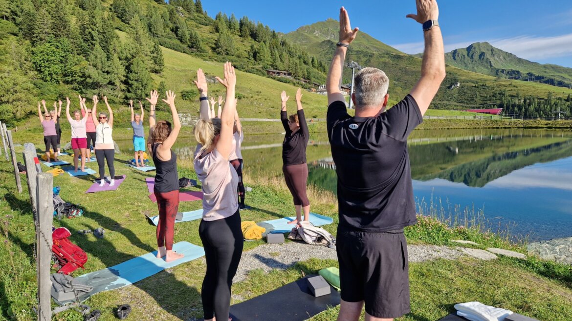 © Rauriser Hochalmbahnen Eine Gruppe macht Yoga im Freien an einem See, umgeben von grünen Hügeln und Bäumen unter strahlend blauem Himmel. (vergrößerte Ansicht)