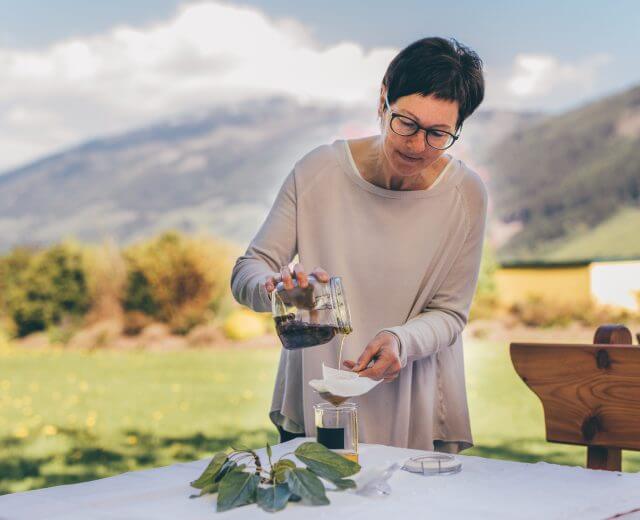 Lungau-SalzburgerLand Tourismus-Michael Groessinger Eine Frau gießt Flüssigkeit durch ein Filterpapier im Freien mit Bergen im Hintergrund.