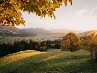 2020_Landschaft_Herbst_©Christian Schartner (174) Sonnenaufgang über einem grünen Tal mit Bäumen, Bergen, Kulinarik und einem fernen, von Blättern umrahmten Dorf.