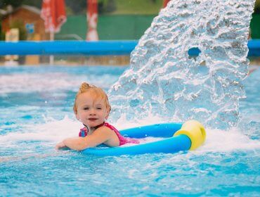 Lächelndes Kleinkind in einem Schwimmbecken in der Nähe eines Wasserbrunnens bei Familienthermen, das den sonnigen Tag genießt.