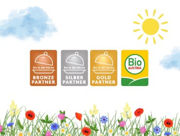 Vier Bio in der Küche-Partnerplaketten und ein Bio Austria-Logo über einem Feld mit Wildblumen, Wolken und einer Sonne.