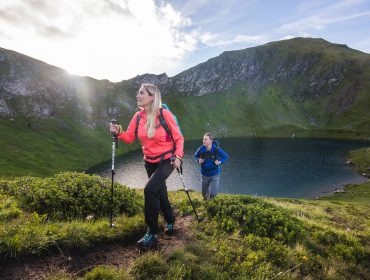 Zwei Frauen, die mit Trekkingstöcken bergauf wandern, umarmen Nachhaltigkeit in der Nähe eines Bergsees unter einem teilweise bewölkten Himmel.