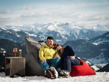 Ein Paar genießt den Sonnenskilauf, entspannt sich auf Sitzsäcken im Schnee mit Getränken und Bergblick.
