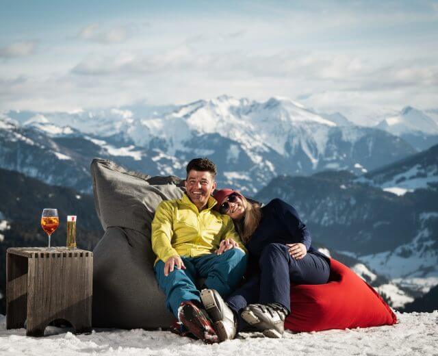Sonnenskilauf bei der Steinbockalm Ein Paar genießt den Sonnenskilauf, entspannt sich auf Sitzsäcken im Schnee mit Getränken und Bergblick.