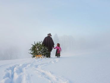 Ein Erwachsener und ein Kind ziehen einen kleinen Weihnachtsbaum auf einem Schlitten durch eine verschneite, neblige Landschaft.
