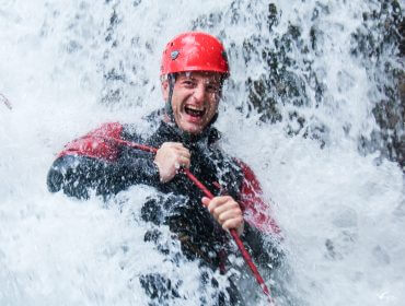 Mann mit rotem Helm und Neoprenanzug unterm Wasserfall