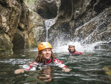 Zwei Personen mit Helmen genießen das Wildwasserschwimmen in einem felsigen Canyon-Fluss in der Nähe eines kleinen Wasserfalls und sehen begeistert aus.