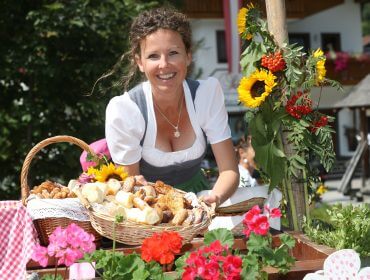 Eine lächelnde Frau präsentiert bei der Eröffnung einen Korb mit Brot und Gebäck, umgeben von bunten Blumen.
