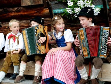 Bauernherbst – Kinder musizieren Drei Kinder in traditioneller Kleidung spielen Akkordeon, während ein Mädchen im Dirndl zwischen ihnen sitzt und lächelt.