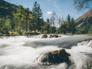 Ein schnell fließender Fluss, still oder prickelnd, schlängelt sich unter blauem Himmel durch eine üppige Berglandschaft.