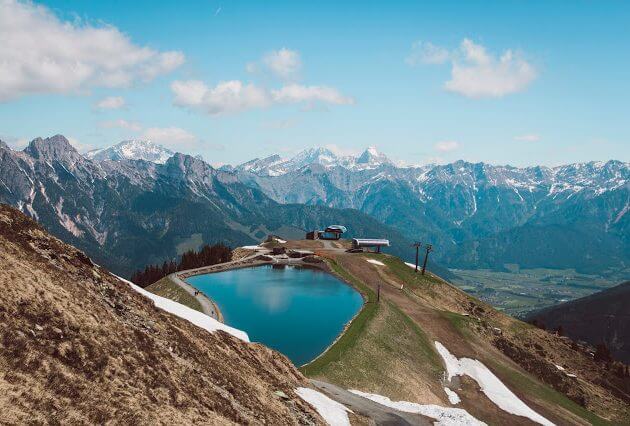 Ein Bergteich mit einer Skiliftstation, umgeben von schneebedeckten Alpen unter blauem Himmel.