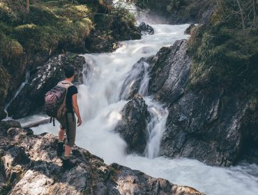 Eine Person mit einem Rucksack steht auf einem Felsen und bewundert einen stillen Wasserfall in einem üppigen Wald.