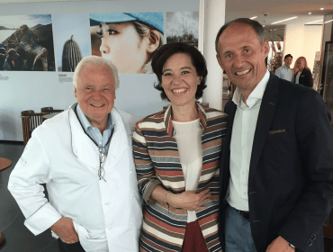 Drei lächelnde Erwachsene posieren zusammen in einem Haus; einer trägt einen weißen Kittel, die anderen tragen Geschäftskleidung.