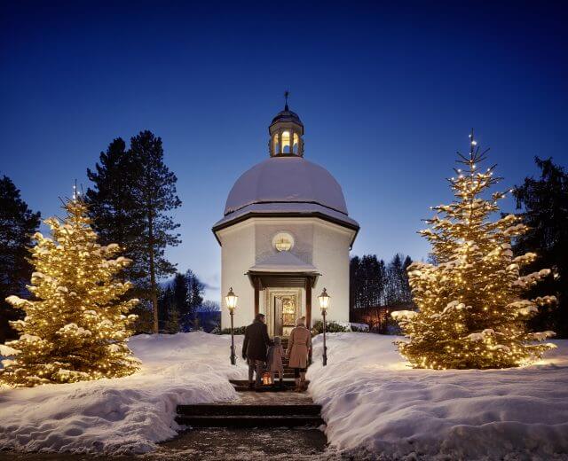 Zwei Personen betreten in der Abenddämmerung eine schneebedeckte Kapelle, flankiert von beleuchteten Weihnachtsbäumen und Laternen.
