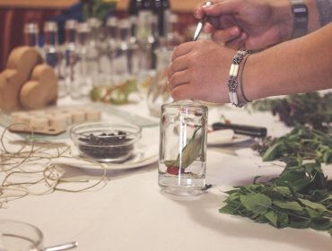 Eine Mitarbeiterin des Löckerwirts füllt Kräuter in eine Glasflasche und stellt sie neben frische Pflanzen und Küchenartikel.