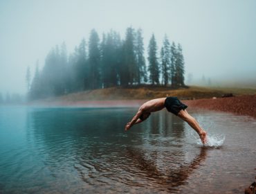 An einem nebligen Instaweekend-Morgen taucht eine Person in einen ruhigen See in der Nähe eines bewaldeten Ufers.