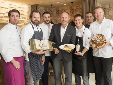 Neun Männer, darunter auch Köche in Uniform, posieren gemeinsam; einer hält einen Teller, ein anderer ein Brot und ein weiterer ein Buch.