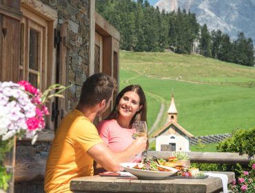 Ein Paar speist im Freien auf einer Holzterrasse mit üppiger grüner Landschaft und atemberaubendem Blick auf den Hochkönig.