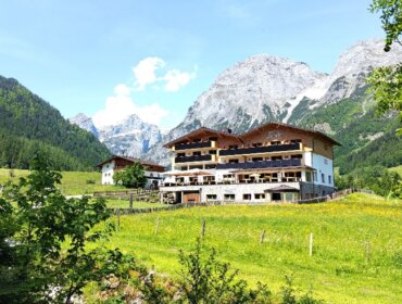 Ein großes Gebäude im Chalet-Stil auf einer grünen Wiese, umgeben von bewaldeten Hügeln und hohen, felsigen Bergen.