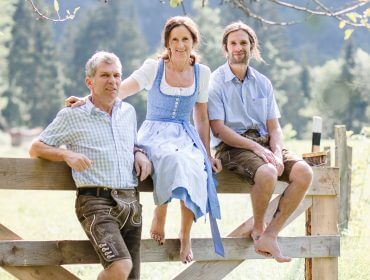 Drei Personen in traditioneller österreichischer Kleidung sitzen barfuß auf einem Holzzaun in einer sonnigen, ländlichen Umgebung im Freien.