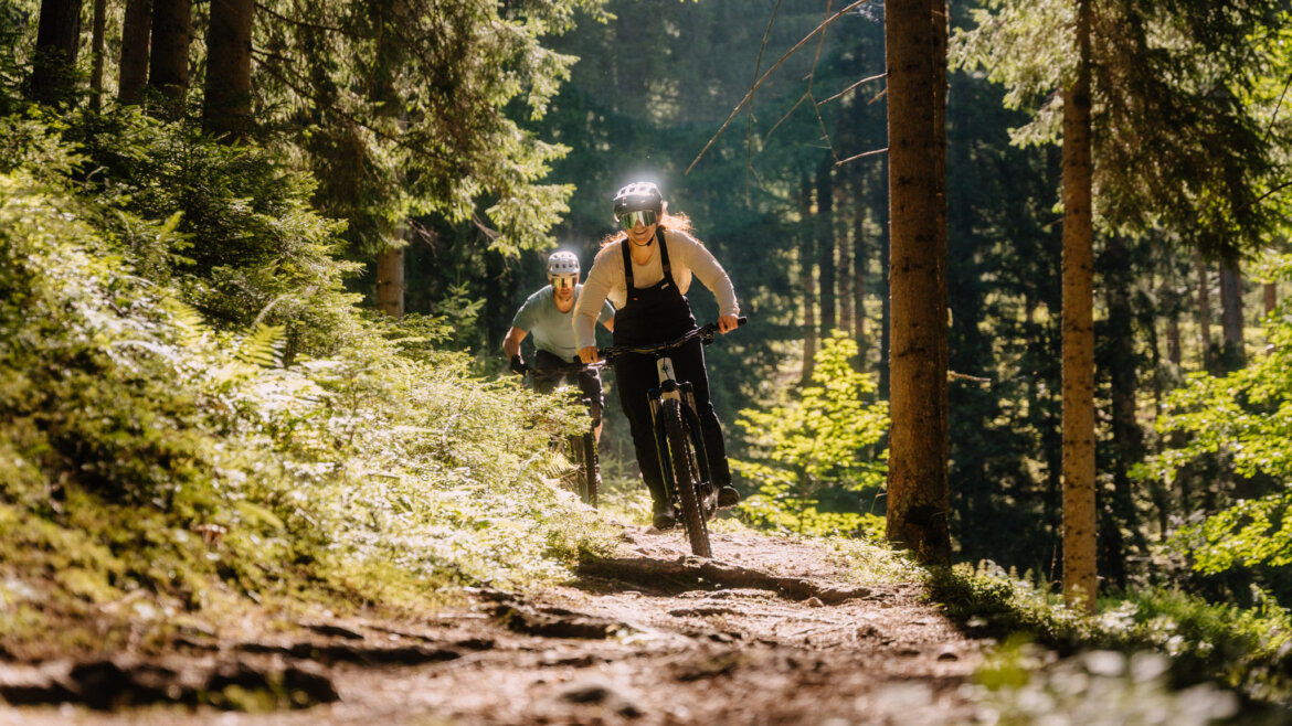 Zwei Menschen genießen das (E-)Mountainbiken in Flachau und fahren einen sonnendurchfluteten Waldweg hinunter, der von üppigen Bäumen gesäumt ist.