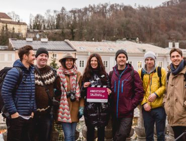 Sieben Entdecker SalzburgerLand posieren im Freien in Winterkleidung mit einem #NEXTSTOP SALZBURG-Schild; dahinter Hügel.
