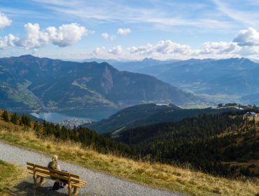 Eine Person sitzt auf dem Bankerl und genießt die grandiose Aussicht über die Hügel, Berge und den Zeller See