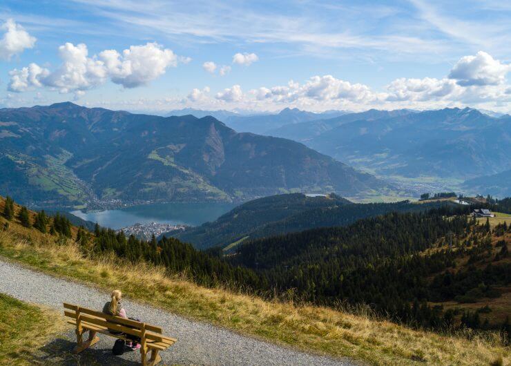 Eine Person sitzt auf dem Bankerl und genießt die grandiose Aussicht über die Hügel, Berge und den Zeller See