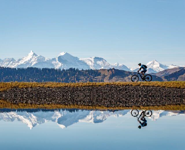 Ein Mountainbike-Fahrer fährt an einem ruhigen See entlang, schneebedeckte Gipfel spiegeln sich im Wasser.