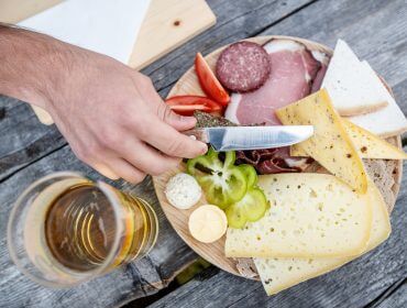 Eine Person schneidet Käse auf einer Holzplatte in Großarltal, mit Fleisch, Gemüse und Käse, neben einem Glas Bier.