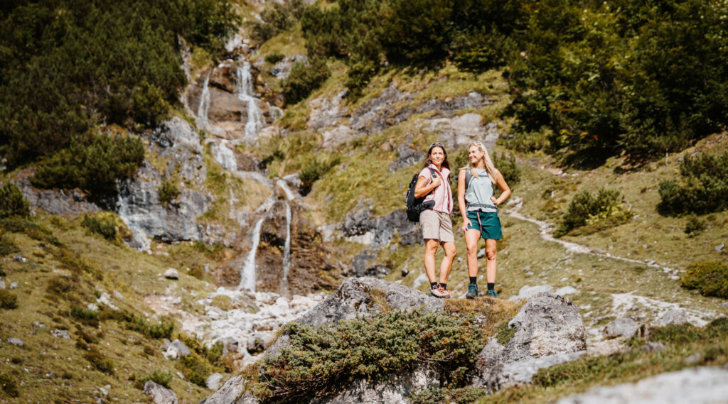 Zwei Frauen stehen auf Felsen an einem Wasserfall und genießen das Kräuterwandern und die Berglandschaft.