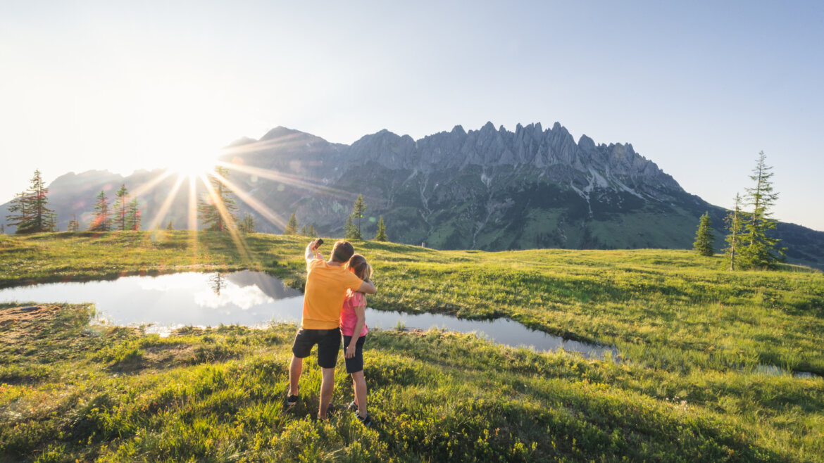 Zwei Menschen umarmen sich an einem Teich auf einer Wiese und genießen die Sonne und die Berge bei ihrem Kräuterwandern.