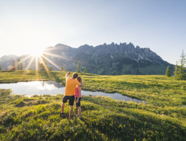Hochkönig Tourismus GmbH Hüttenhüpfer Tour Zwei Menschen umarmen sich an einem Teich auf einer Wiese und genießen die Sonne und die Berge bei ihrem Kräuterwandern.