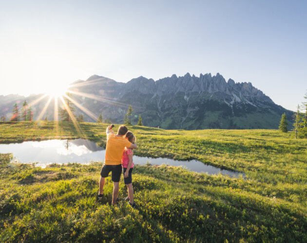 Hochkönig Tourismus GmbH Hüttenhüpfer Tour Zwei Menschen umarmen sich an einem Teich auf einer Wiese und genießen die Sonne und die Berge bei ihrem Kräuterwandern.
