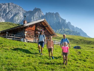 Eine Familie genießt das Familienwandern auf einem grasbewachsenen Hügel in der Nähe einer Holzhütte mit Bergen im Hintergrund.