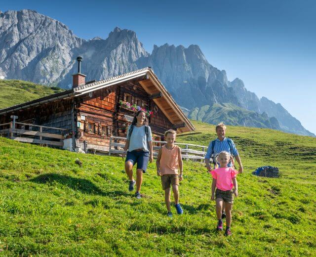 Eine Familie genießt das Familienwandern auf einem grasbewachsenen Hügel in der Nähe einer Holzhütte mit Bergen im Hintergrund.