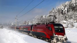 Bahn_öbb_Zug_Railjet_Anreise Ein roter Personenzug fährt unter strahlend blauem Himmel durch eine verschneite Landschaft am Rande eines Waldes.