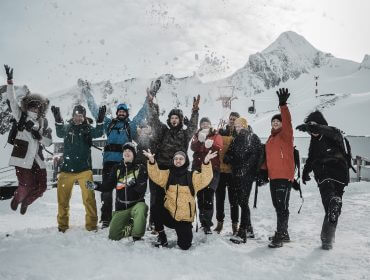 ZASK_Instaweekend_Gruppenfoto Edit-2 Eine Gruppe in Winterkleidung lacht und wirft mit Schnee, im Hintergrund die verschneiten Berge von Zell am See Kaprun.