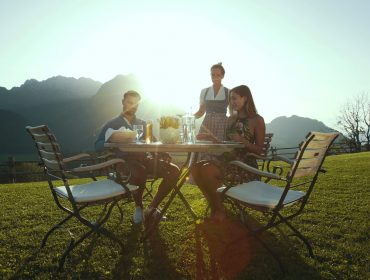 Drei Personen, die eine Mahlzeit im Freien an einem Tisch auf einem grasbewachsenen Rasen mit Bergen und Sonne im Hintergrund genießen.