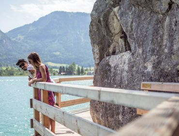 2017-08-02-SLT-Roadtrip-Tag Zwei Personen lehnen an einem Holzgeländer und genießen den stillen oder prickelnden See mit den Bergen im Hintergrund.