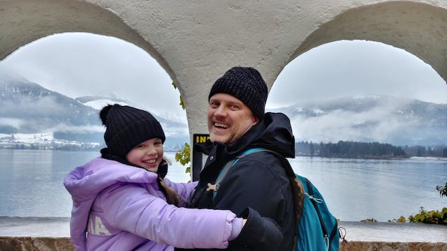 Ein Mann und ein Mädchen in Winterkleidung lächeln durch einen Torbogen mit einem See und verschneiten Bergen im Hintergrund.