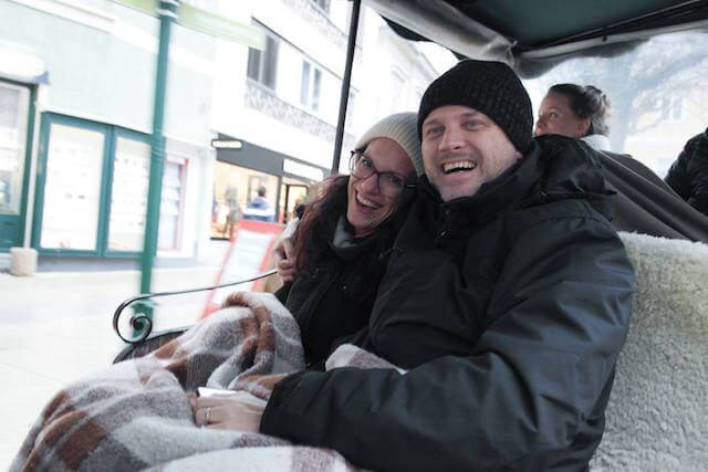 Ein lächelndes, winterlich gekleidetes Paar sitzt bei einer Kutschfahrt eng aneinander gedrängt in einer Decke.