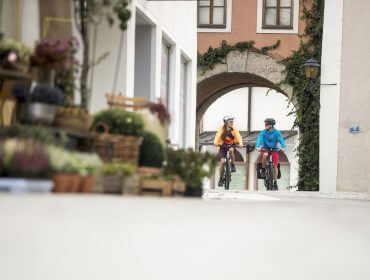 Zwei Personen fahren mit dem Fahrrad den Tauernradweg entlang, vorbei an einer malerischen, blumengesäumten Straße und einem gewölbten Tor.