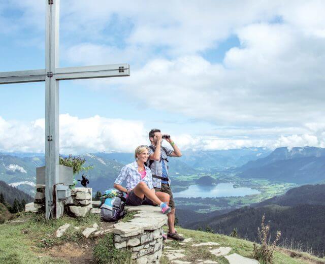 Wandern-Zwo¦êlferhorn(1) Zwei Pilgern-Wanderer rasten an einem Gipfelkreuz mit Blick auf ein malerisches Tal mit See und Bergen.