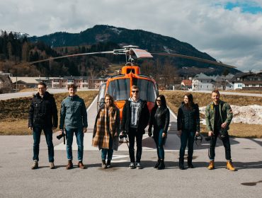 Sechs Personen stehen vor einem orangefarbenen Hubschrauber auf einem Hubschrauberlandeplatz und halten einen myinstaweekend-Moment mit Bergen im Hintergrund fest.