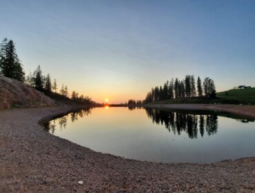 Sonnenuntergang über einem ruhigen See mit Bäumen, die sich im Wasser spiegeln, und einem felsigen, sandigen Ufer im Vordergrund.