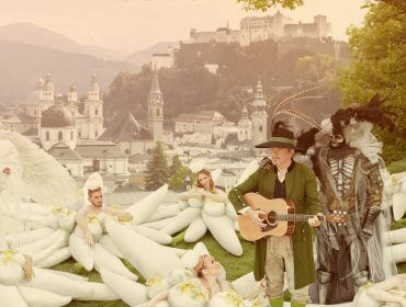 Menschen in skurrilen Life Ball-Kostümen posieren auf einem Hügel mit einer Stadt und einem Schloss im Hintergrund; einer spielt Gitarre.