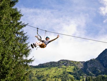 Eine Person, die an einem sonnigen Tag mit dem Ziplining durch die Luft über Saalbachs grüne Berge und Bäume gleitet.