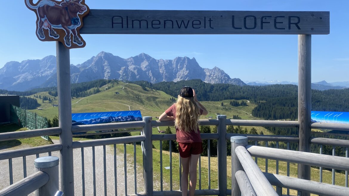 Ein Mädchen steht an einem Schild der „Almenwelt Lofer“ und blickt an einem sonnigen Tag auf die Berge und die grünen Hügel. (vergrößerte Ansicht)